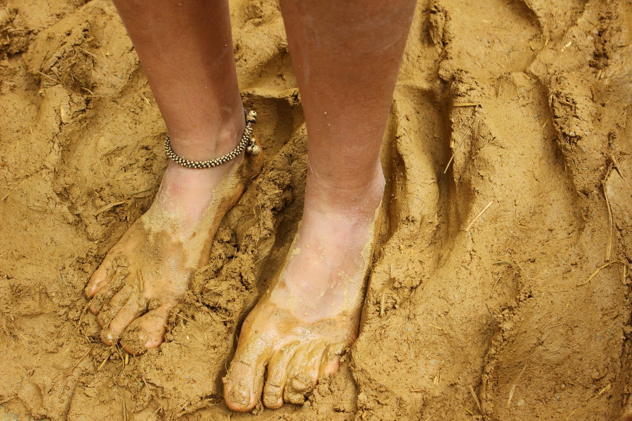 Bare feet coated in wet orange clay treading a cob mix with straw in the mixing pit