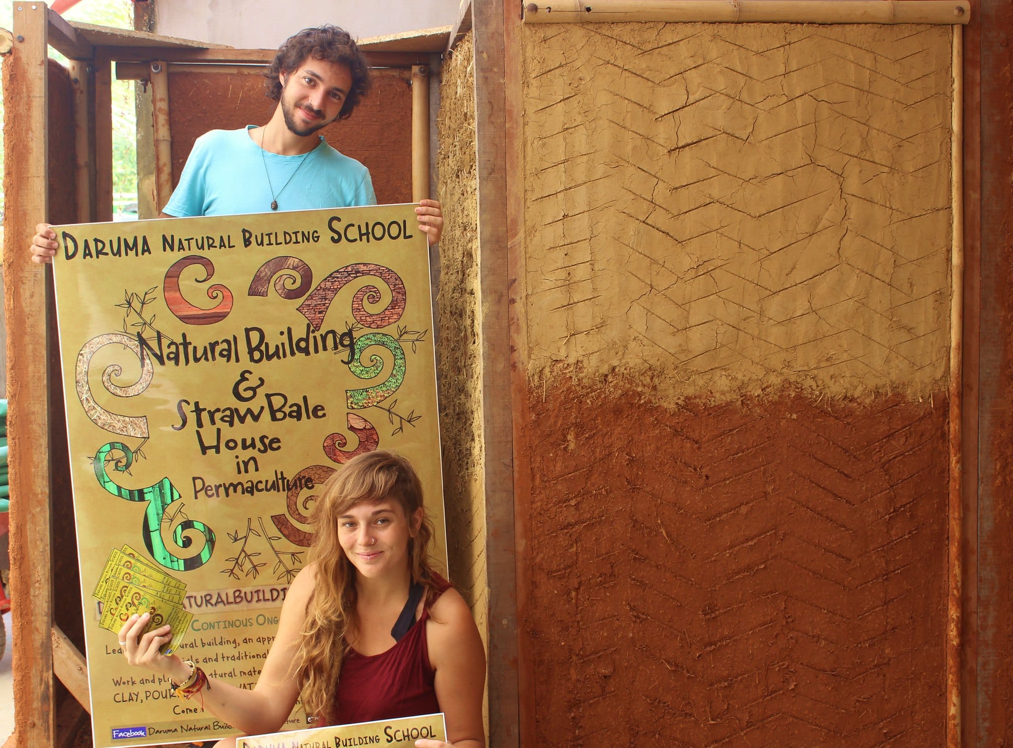 Two smiling Daruma students hold their hand-drawn Natural Building School poster beside an earthen wall.