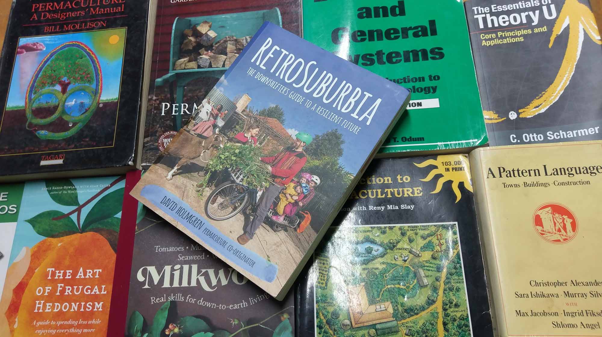 Overhead flat lay of a stack of permaculture and systems theory books including Permaculture: A Designers' Manual, RetroSuburbia and A Pattern Language