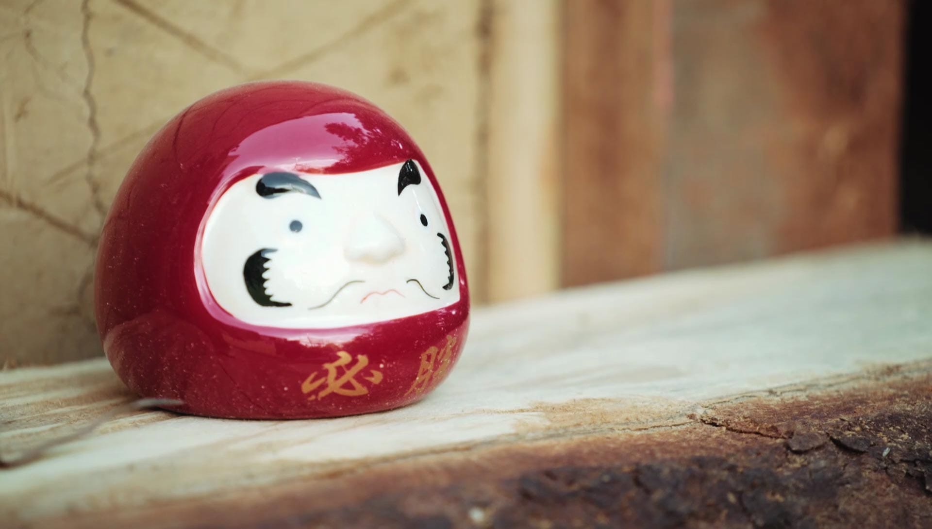 A glossy red ceramic daruma doll with gold Japanese lettering resting on a wooden beam in front of an earthen wall