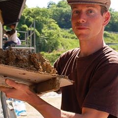 Kyle Holzhueter holding a tray of wet clay plaster, with a rice field and scaffolding behind