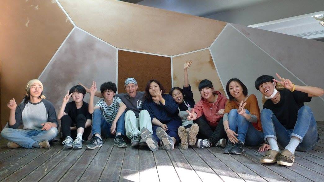 Nine young volunteers posing together on a wooden deck beside an earthen wall at Daruma