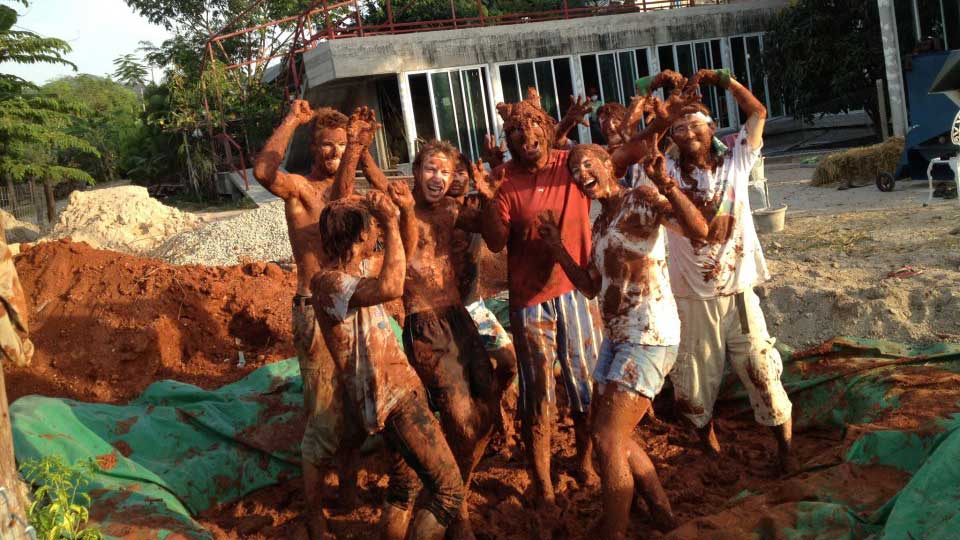 Eight students covered head to toe in mud, cheering with arms raised on a building site