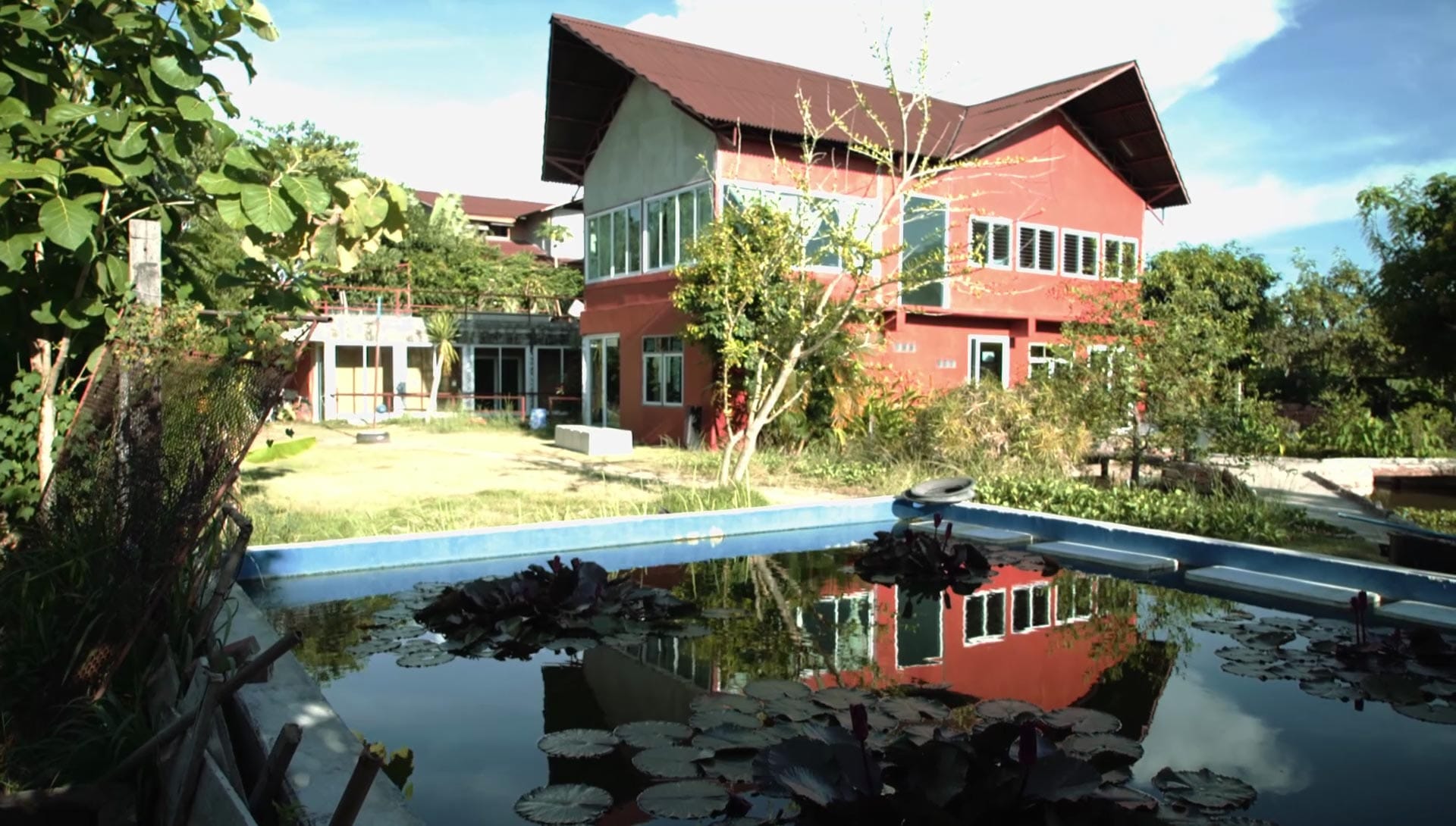 Wide view of the red two-storey schoolhouse at Daruma seen across the lily-pad-covered natural pool, blue-edged deck and lawn in the foreground