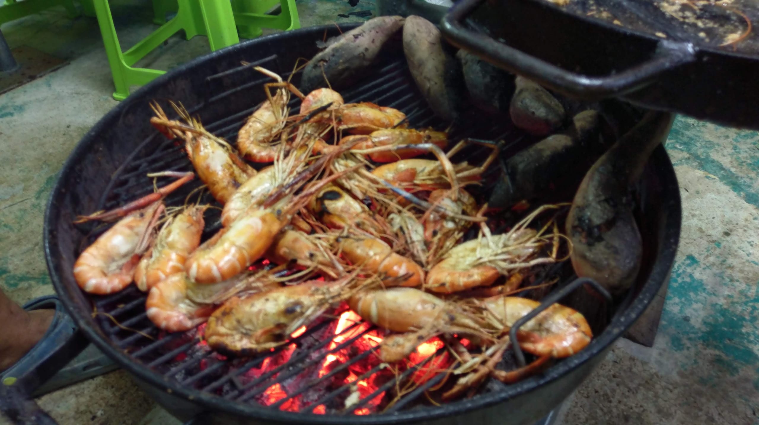 Overhead view of whole grilled prawns with charred shells on a barbecue grill over glowing coals, roasted sweet potatoes alongside