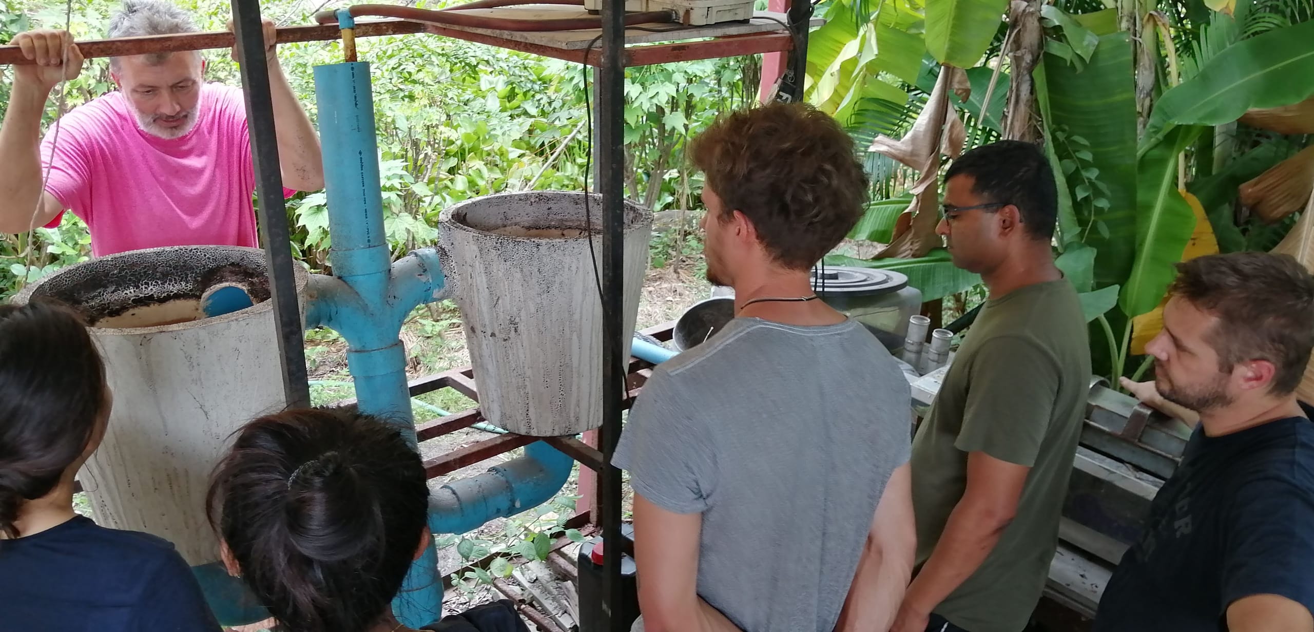 Course students gathered around the natural pool's filtration rig of plant-filter pots and blue PVC piping, with an instructor adjusting the pipework