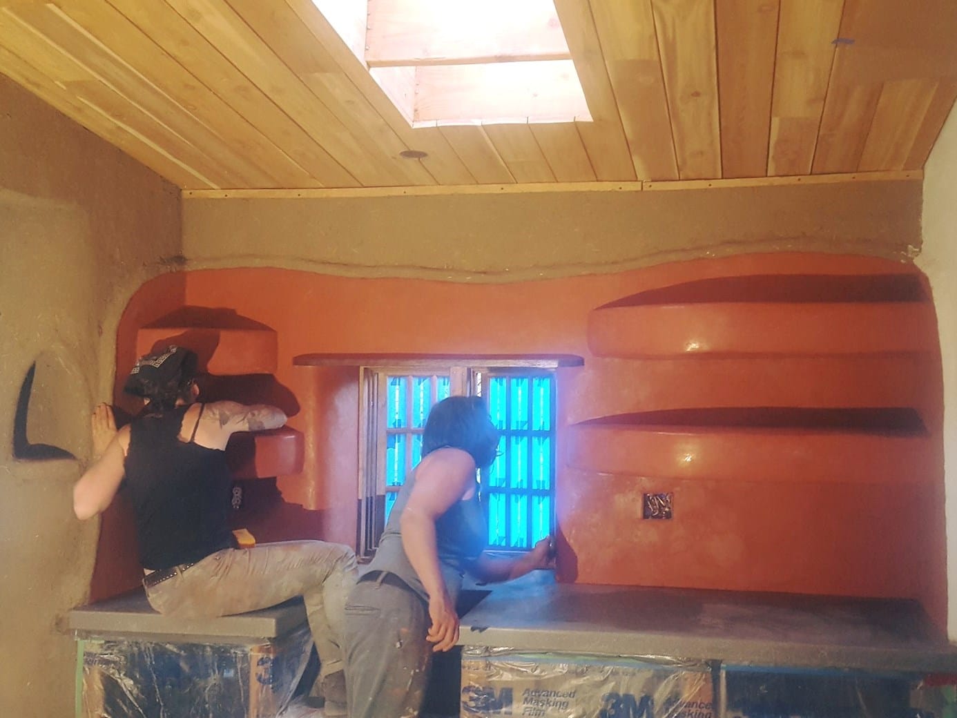 Two people plastering curved earthen wall niches inside a cob building under construction, beside a bright blue-glazed window