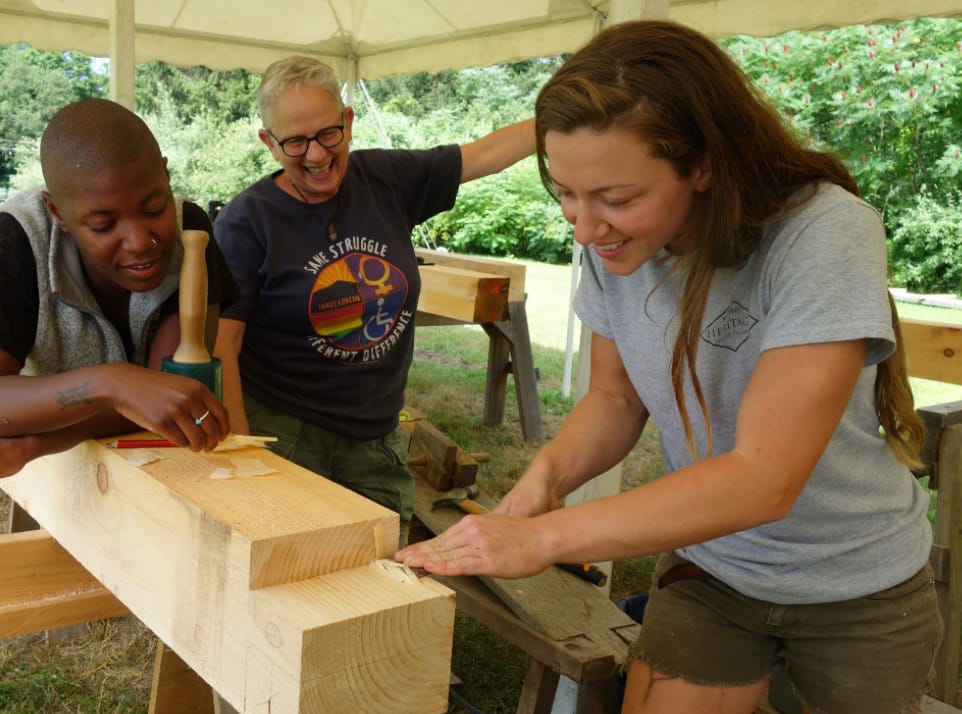 Three women laughing under a canvas tent while working timber-frame joinery with a chisel and mallet, during the Kamimomi workshop
