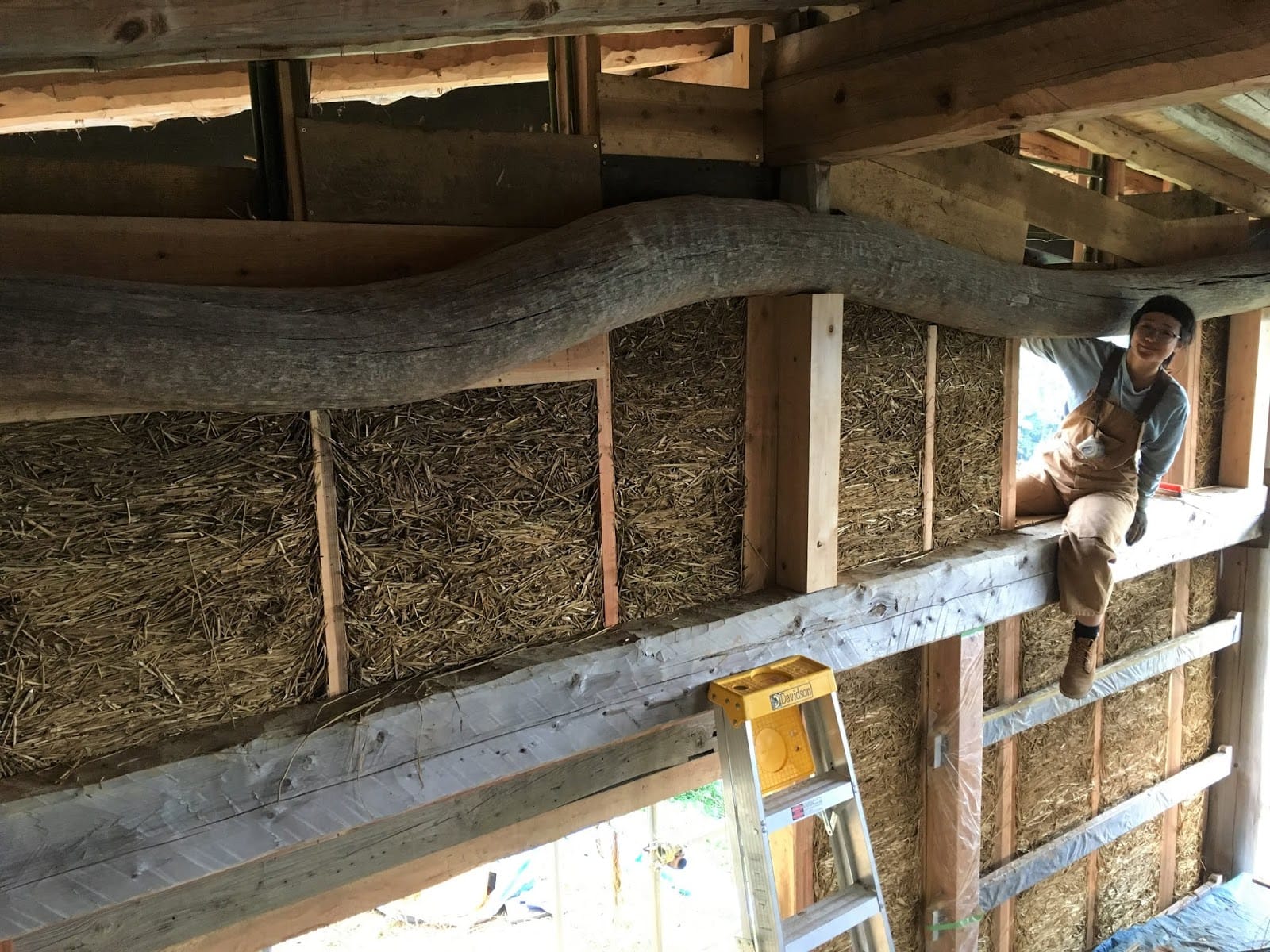 Interior of a straw-bale timber-frame building under construction, showing a curved wooden beam and straw infill panels between the studs