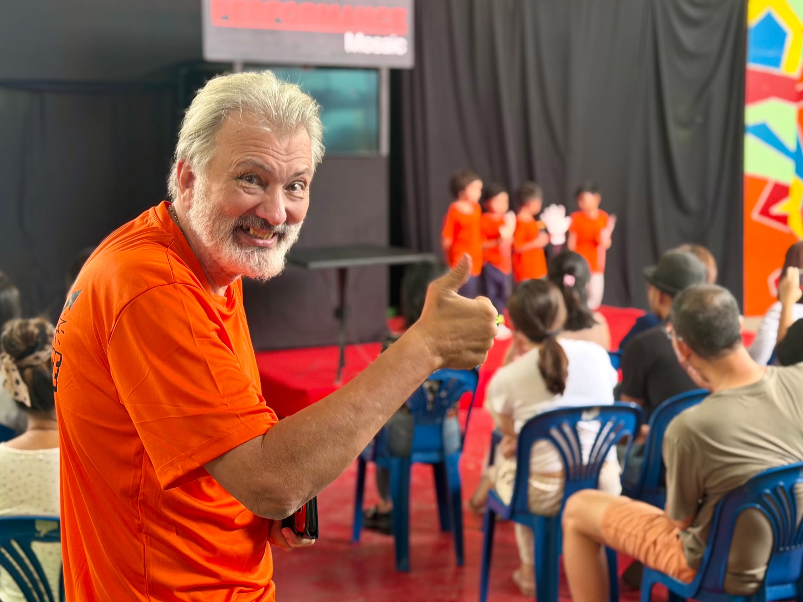 Neil giving a thumbs-up at a Mosaic School performance, with students in costume on stage behind him