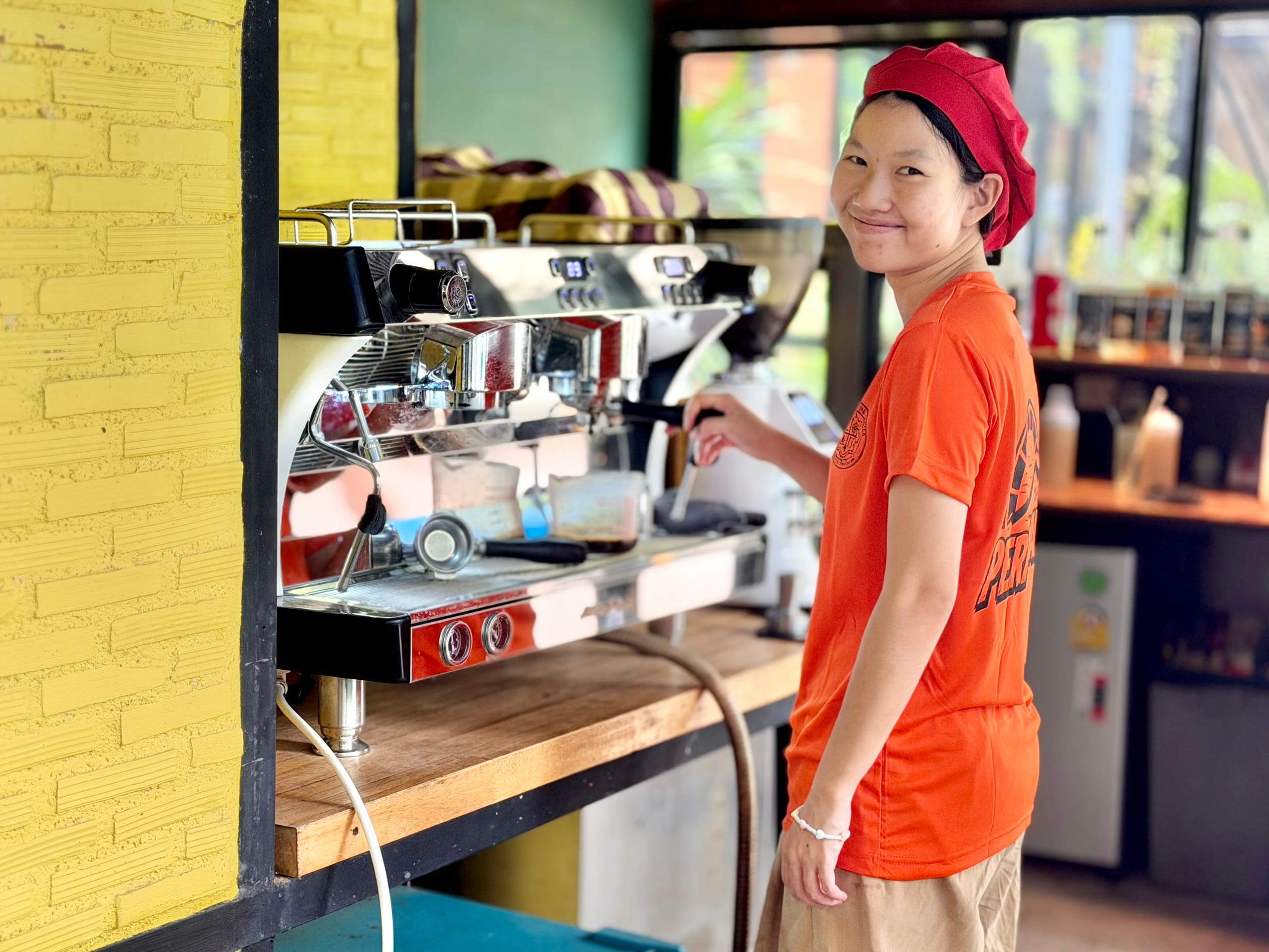 A young woman in a Mosaic-branded staff t-shirt smiling while working the espresso machine in the campus cafe