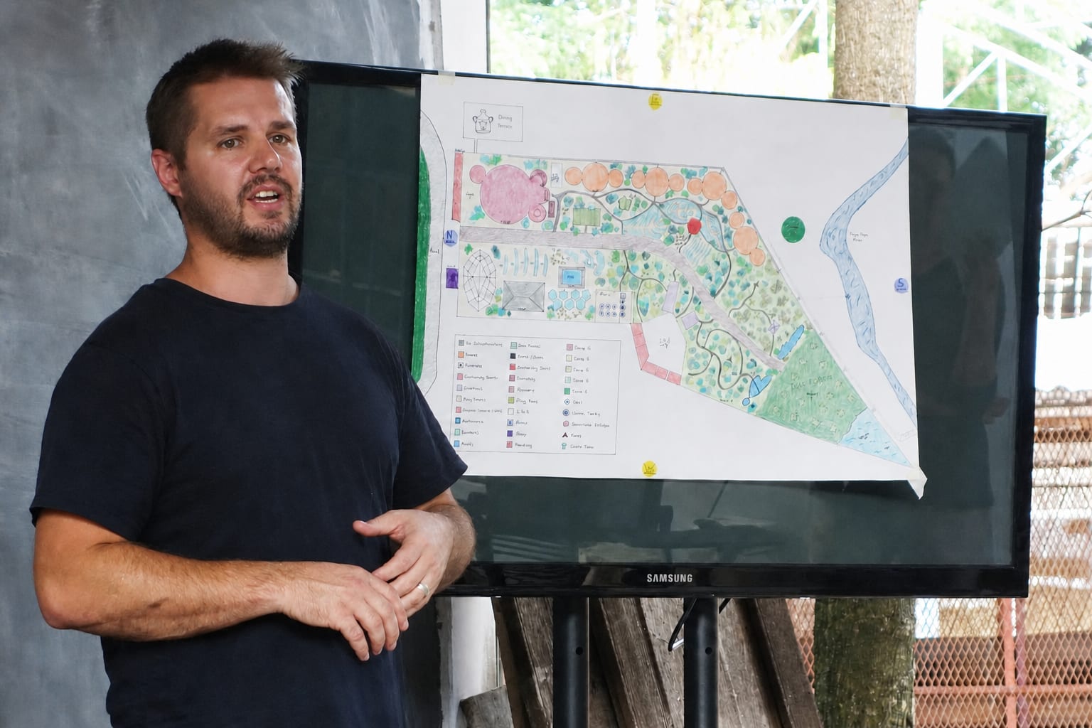 A relaxed permaculture teacher presents a hand-drawn site plan to an audience