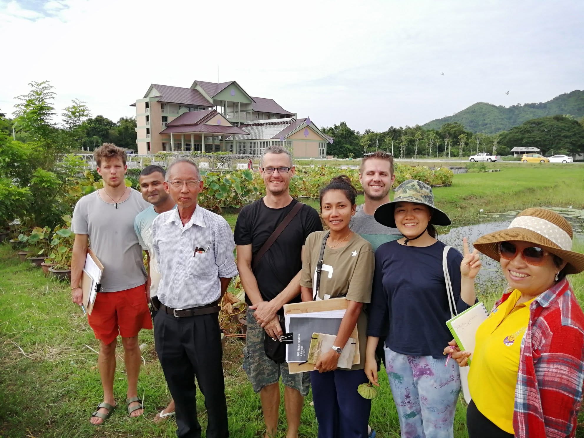 A group of course visitors on a campus tour beside the lotus pond, holding notebooks, with the school building and forested hills behind