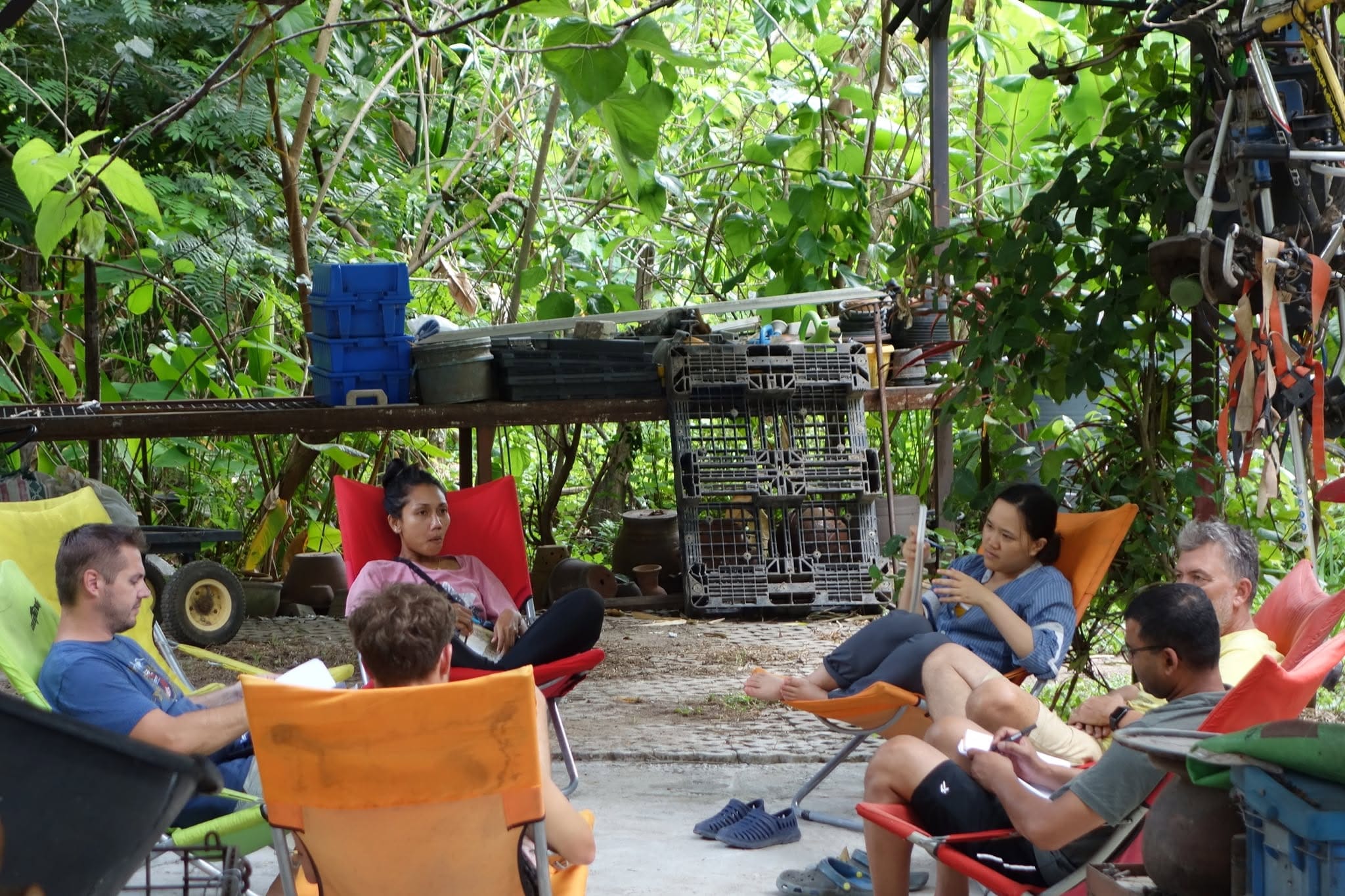 A circle of volunteers and staff in camp chairs with notebooks, talking through the day in a shaded garden