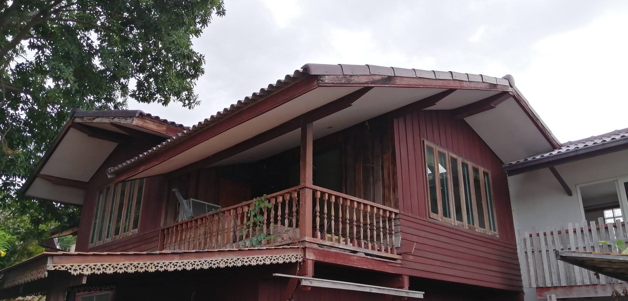 Low-angle view of a traditional two-storey dark red Thai wooden house with a curved tiled roof and turned balcony railings on the Daruma campus.