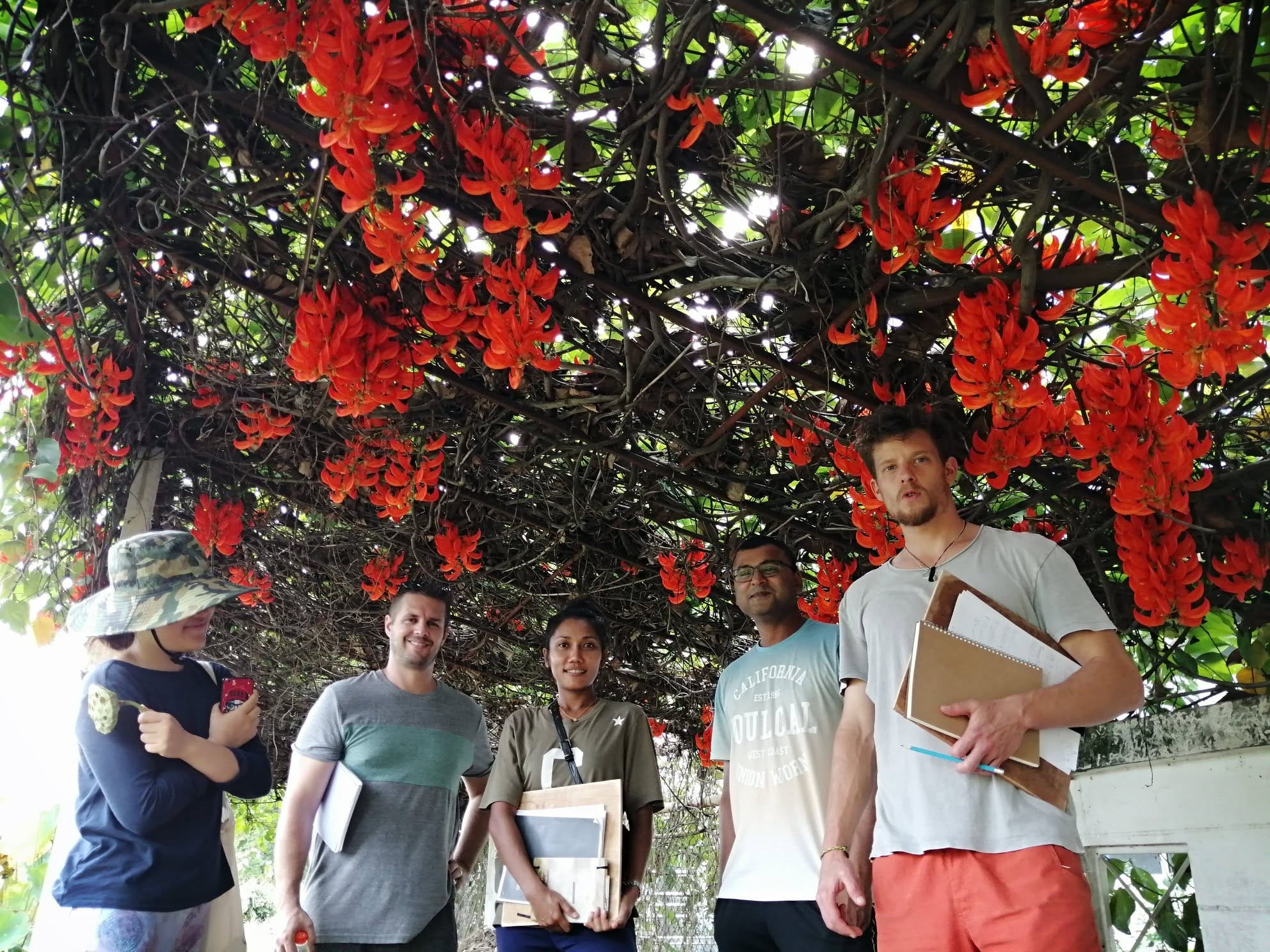Five course students with notebooks and clipboards standing together under a pergola covered in flowering red-orange vine