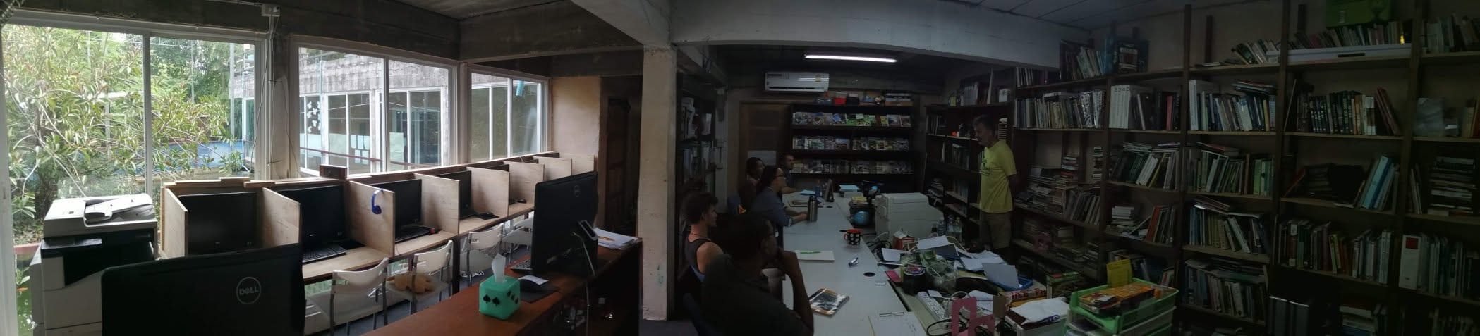 A panoramic view inside the library and computer lab, floor-to-ceiling bookshelves behind a row of desktop workstations and a table of readers by the window