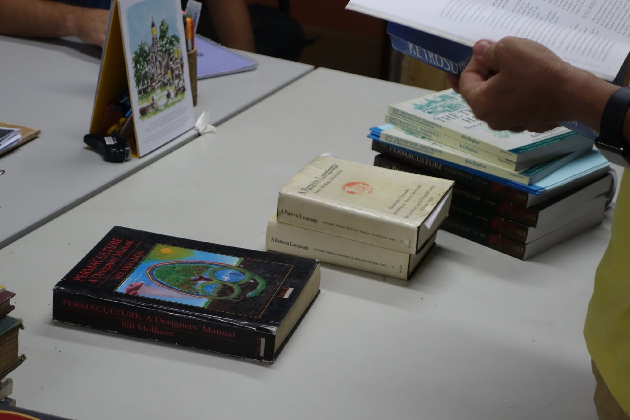 A stack of permaculture reference books on a table, including Bill Mollison's Designers' Manual, two copies of A Pattern Language and several copies of The Transition Handbook