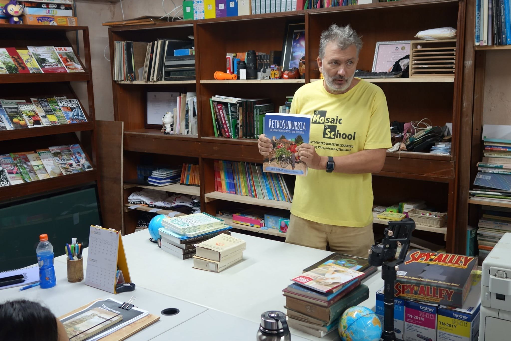 Neil in a Mosaic School t-shirt holding a copy of RetroSuburbia in the office library