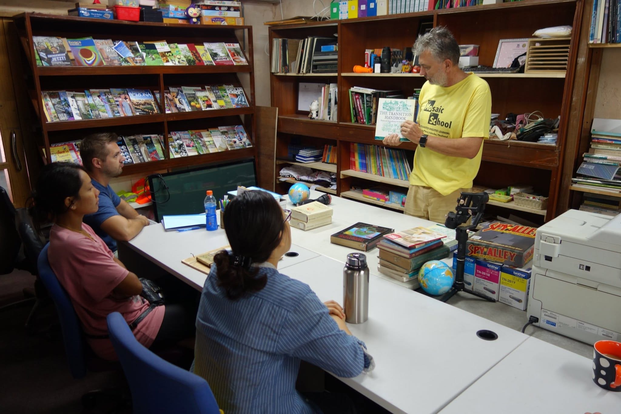 A grey-haired instructor in a yellow Mosaic School t-shirt holds up The Transition Handbook to a group of seated students