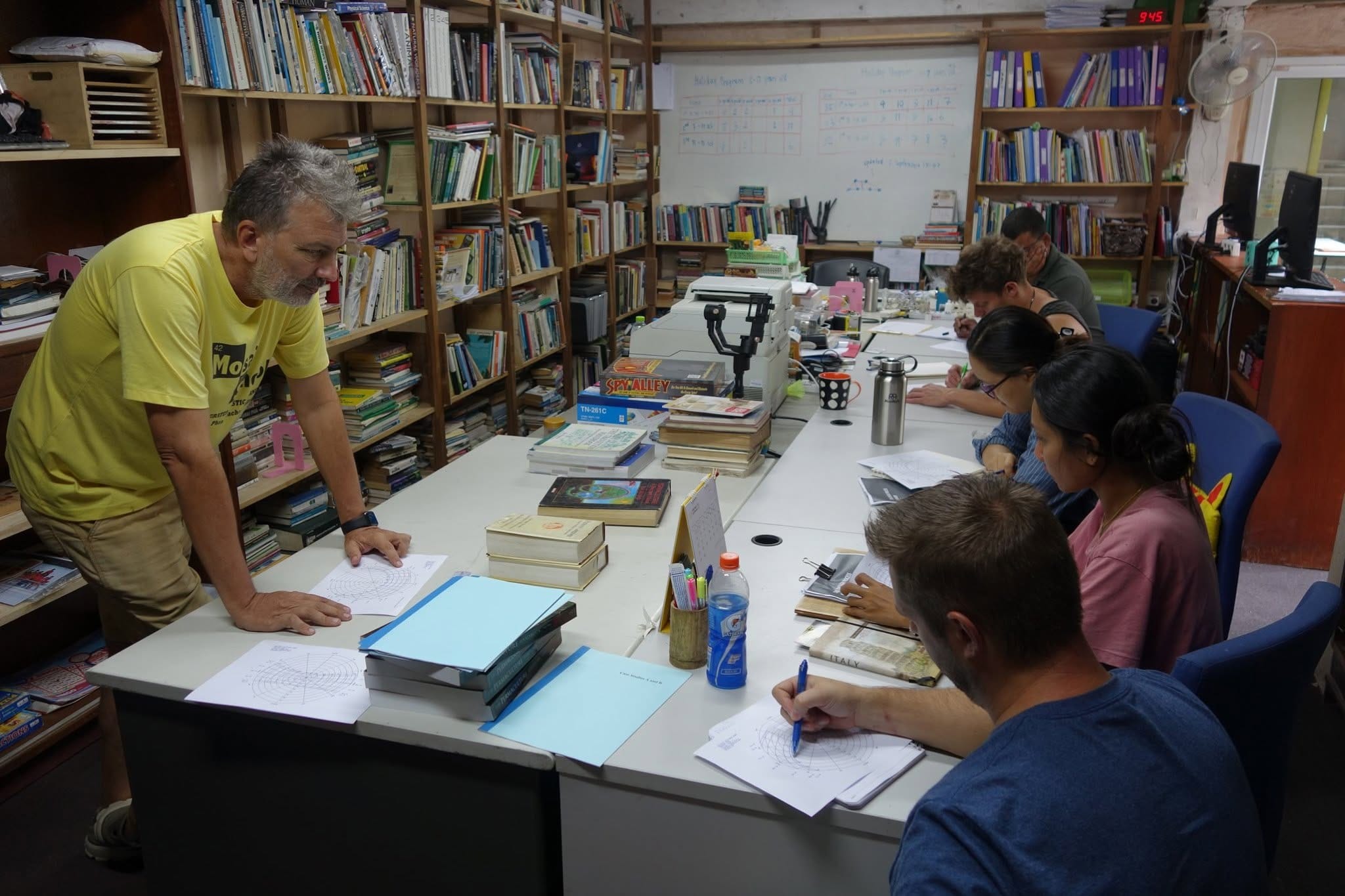 An instructor in a yellow t-shirt leans over a long table to guide a small group working through permaculture design worksheets in a book-lined room.