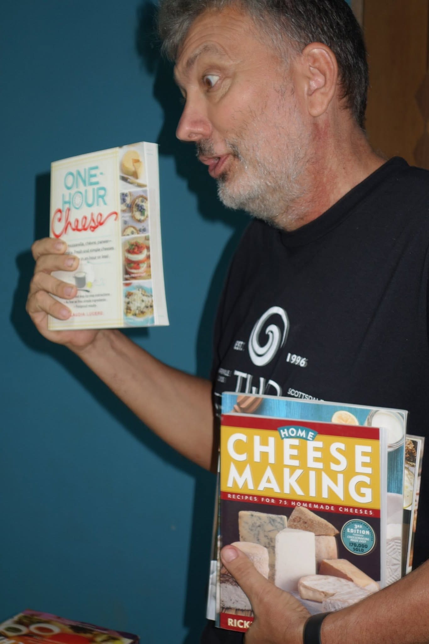 A man in a black t-shirt holds up two cheesemaking reference books, One-Hour Cheese and Home Cheese Making, against a teal wall