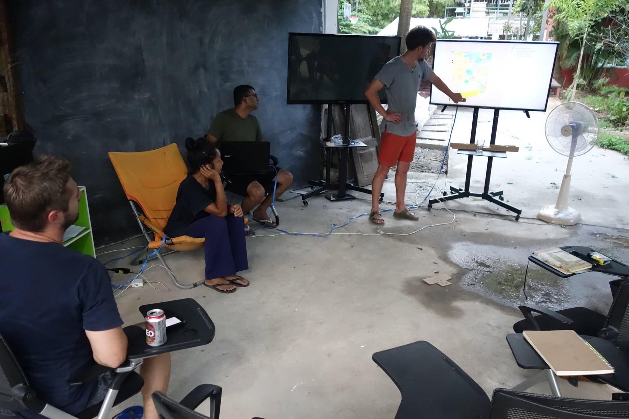 An instructor points at a colourful site plan on a screen while a small seated group watches, with a camp chair in the foreground