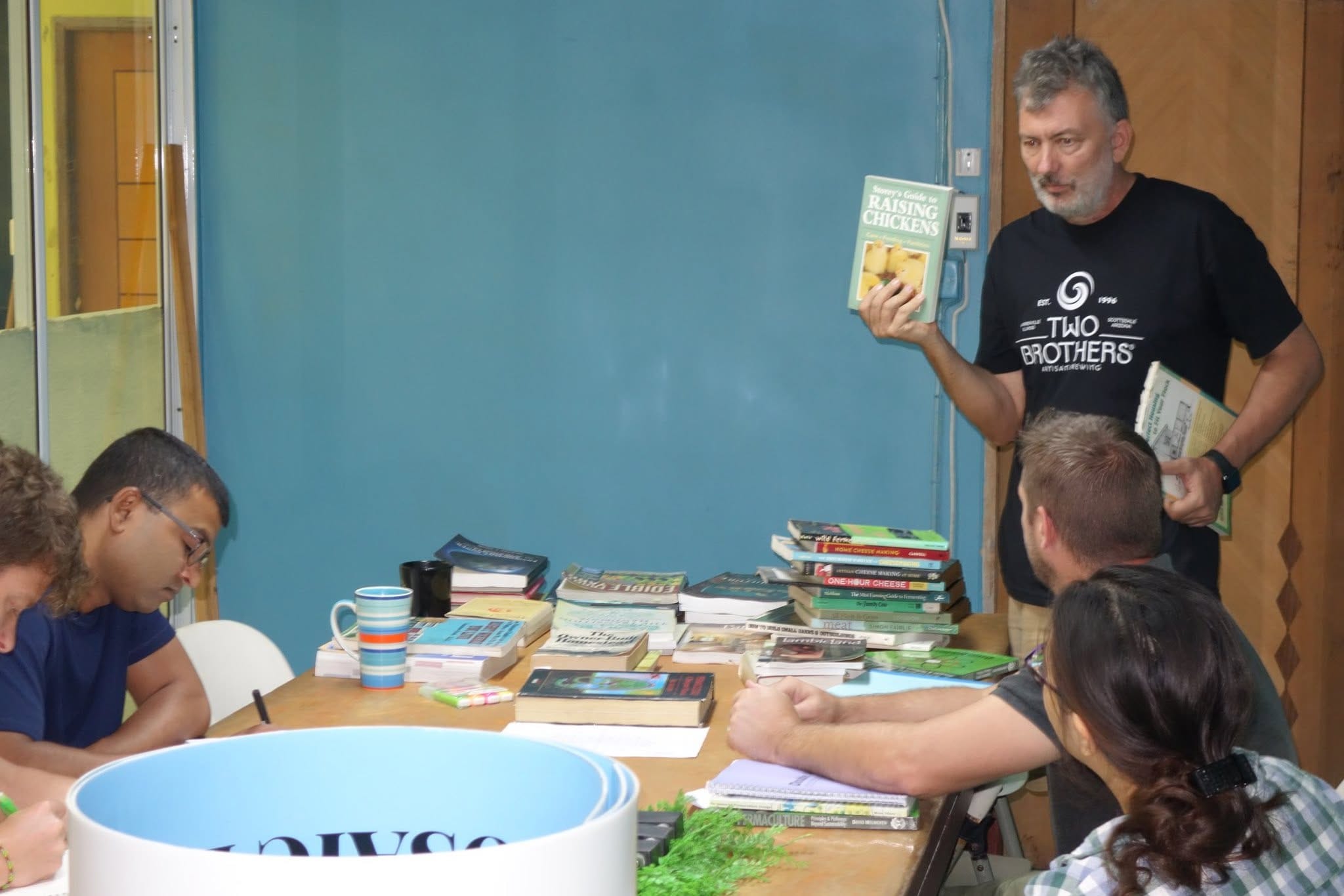 An instructor holding up a guide to raising chickens while students browse stacks of books