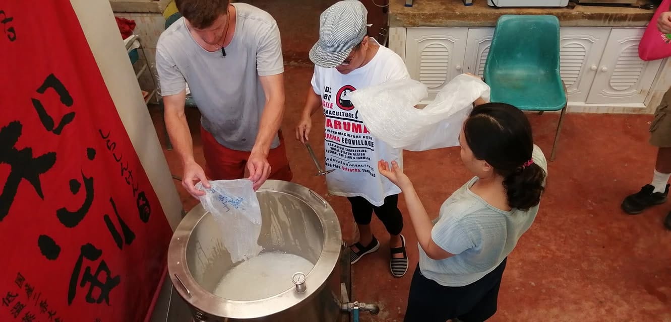 Three people straining fresh milk into a steel pot during cheese making, one in a Daruma t-shirt