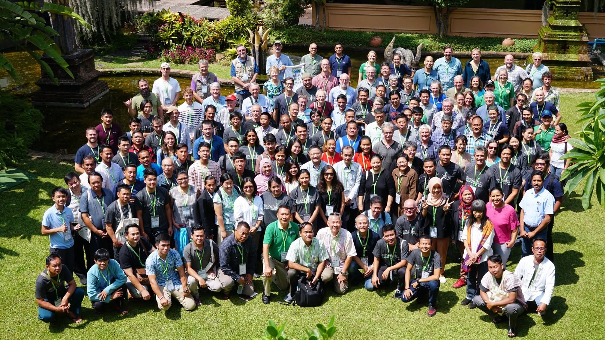An aerial view of a permaculture design gathering, rows of attendees on a lawn beside a pond