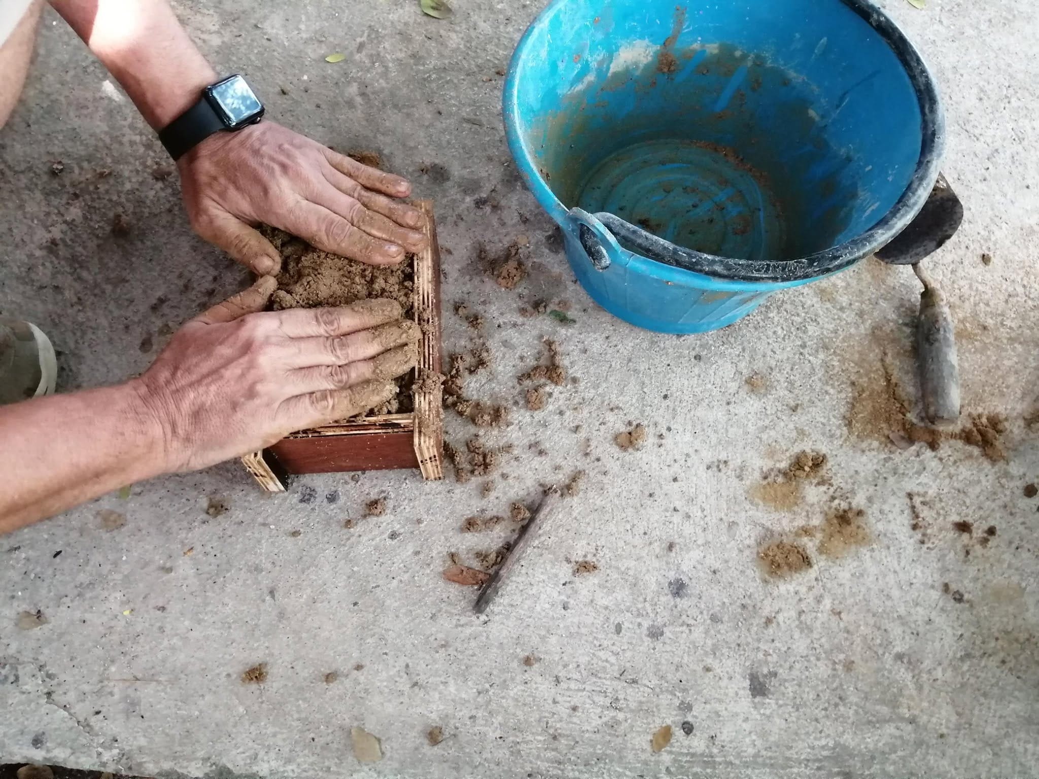 Two muddy hands pressing earthen mix into a wooden brick mold on the ground
