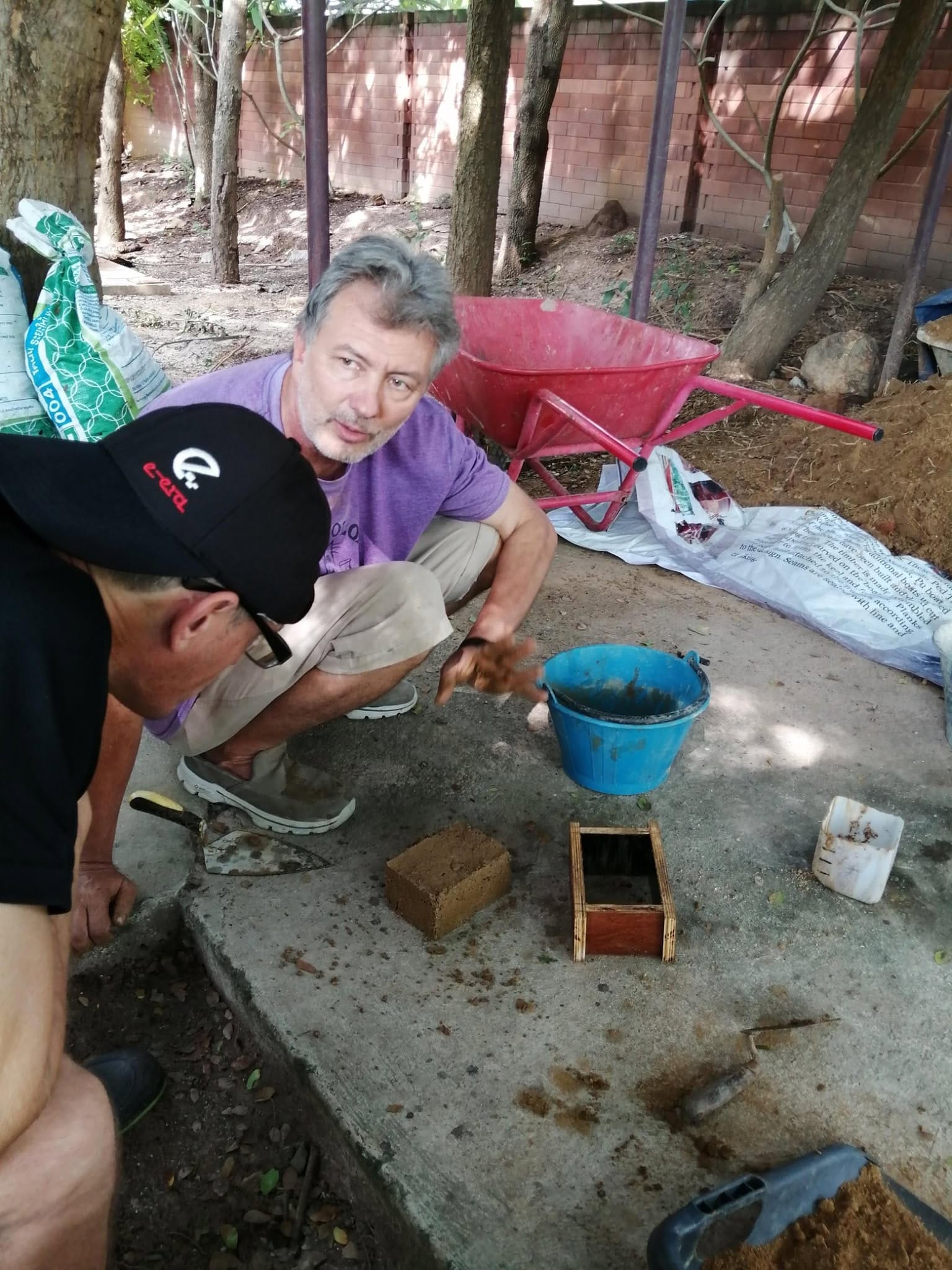 Two students crouching over a freshly made earthen brick and its wooden mold