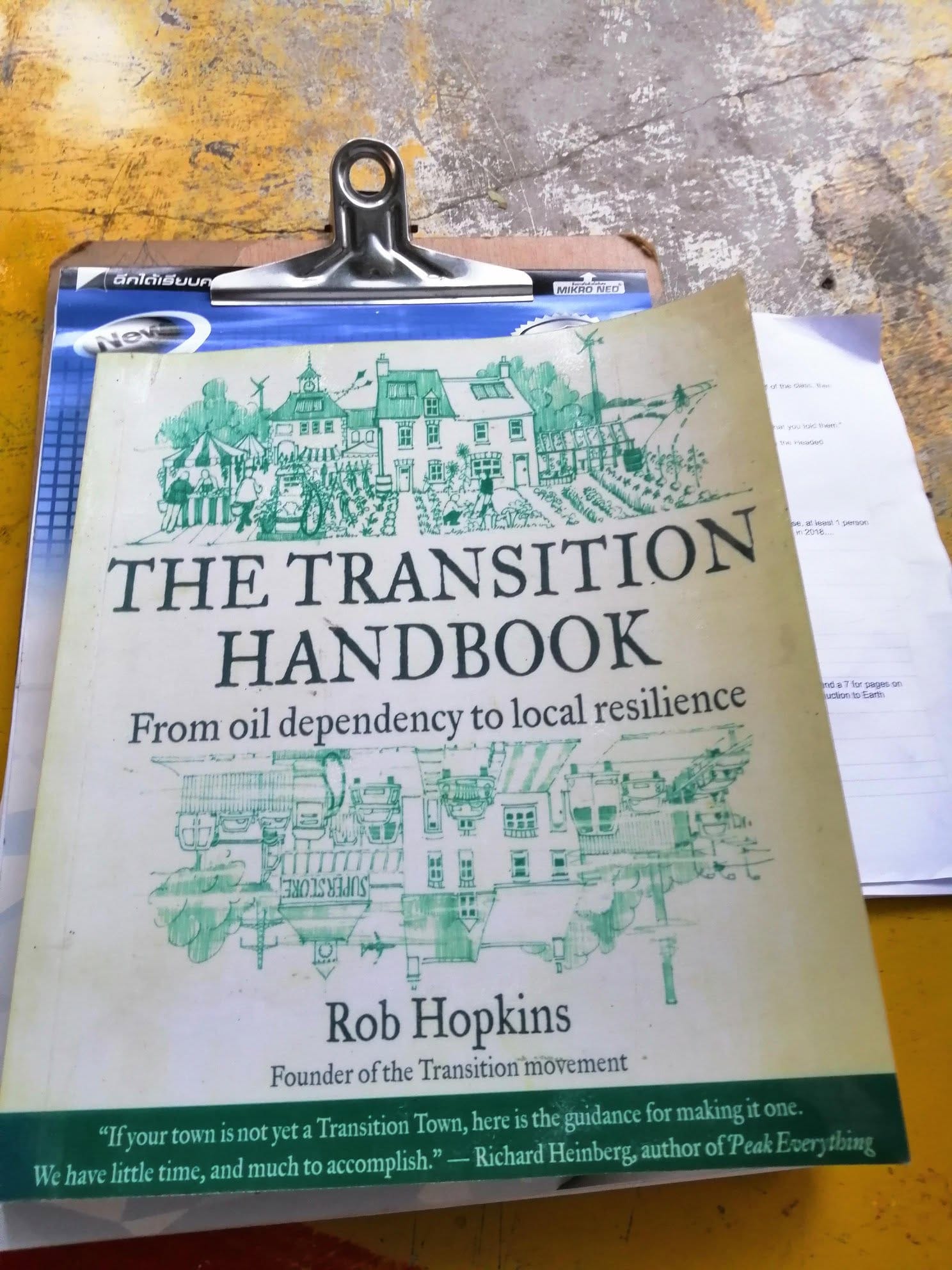 A copy of The Transition Handbook by Rob Hopkins resting on a clipboard on a weathered yellow table, with a printed handout partly visible beside it