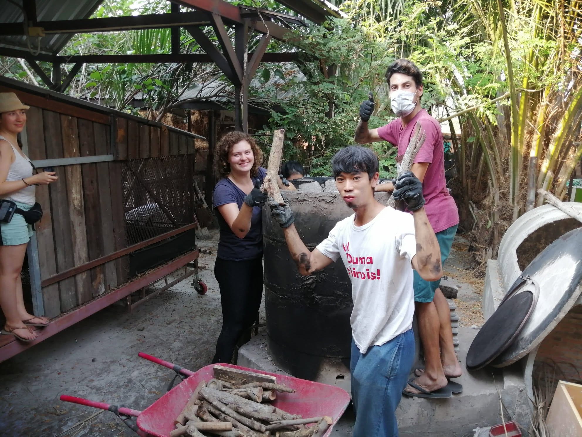 Four muddy-handed volunteers smiling around the biochar kiln at Daruma, holding sticks of wood, one wearing a face mask