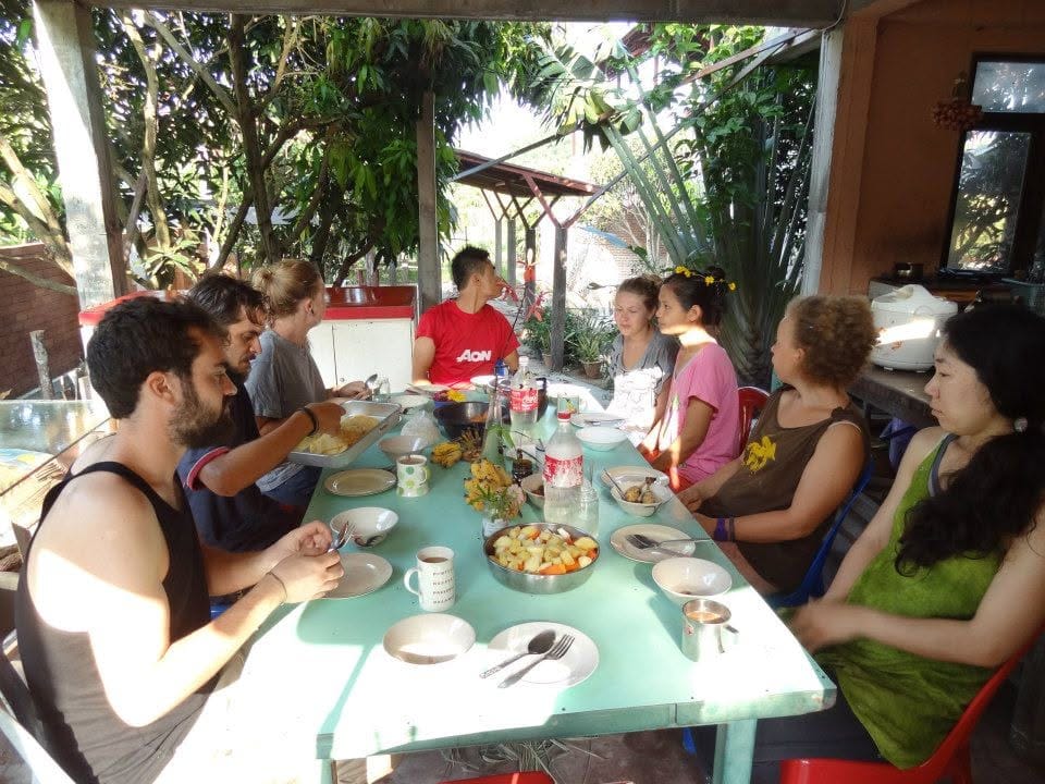 Volunteers and students sharing a communal meal at a long table under a shaded outdoor roof at Daruma