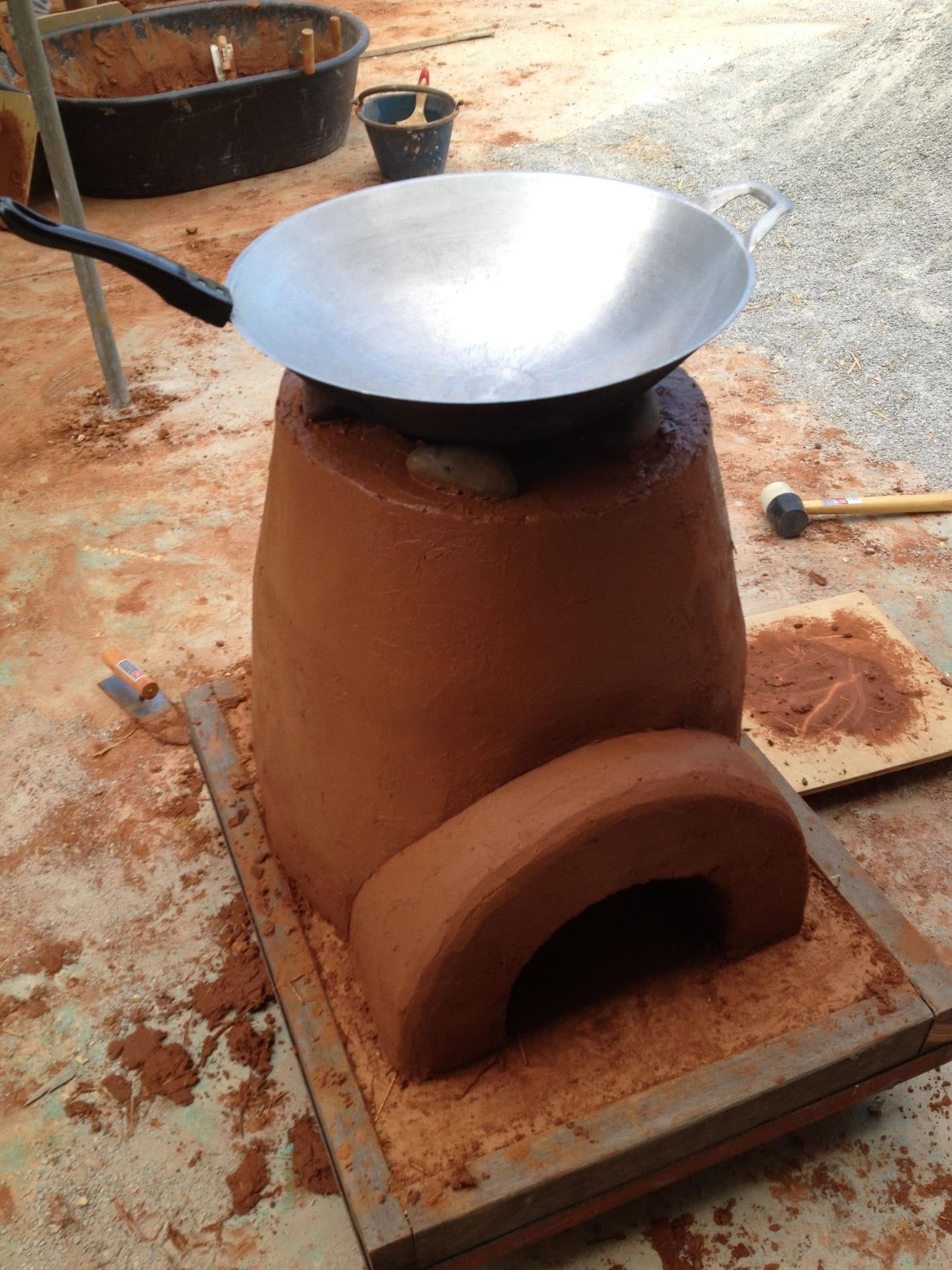 A hand-built reddish-brown cob rocket stove with an arched fuel-feed opening, topped with a steel wok, surrounded by mixing tubs and hand tools at Daruma