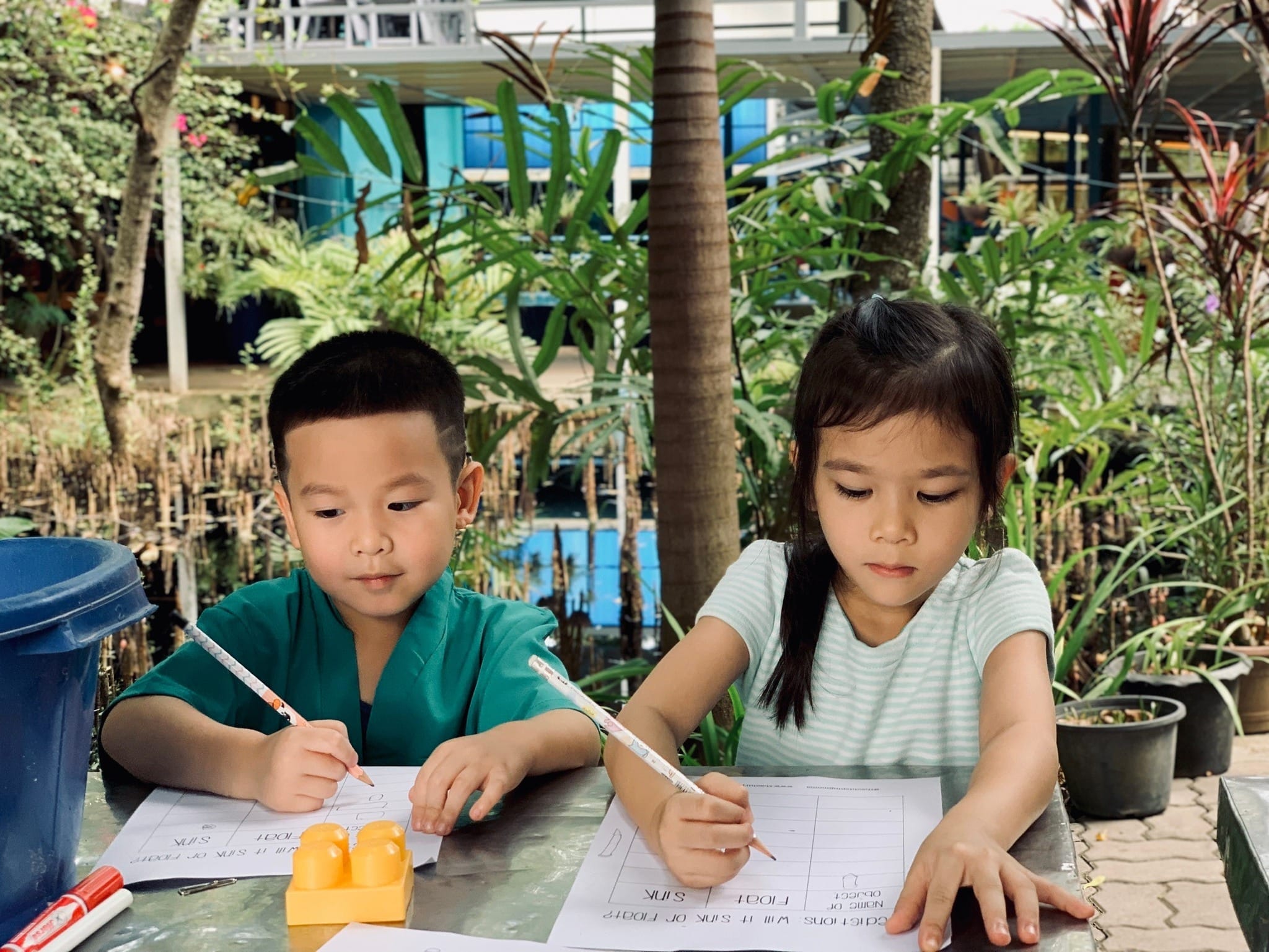 Two young Mosaic School students writing on science worksheets at an outdoor table surrounded by tropical greenery