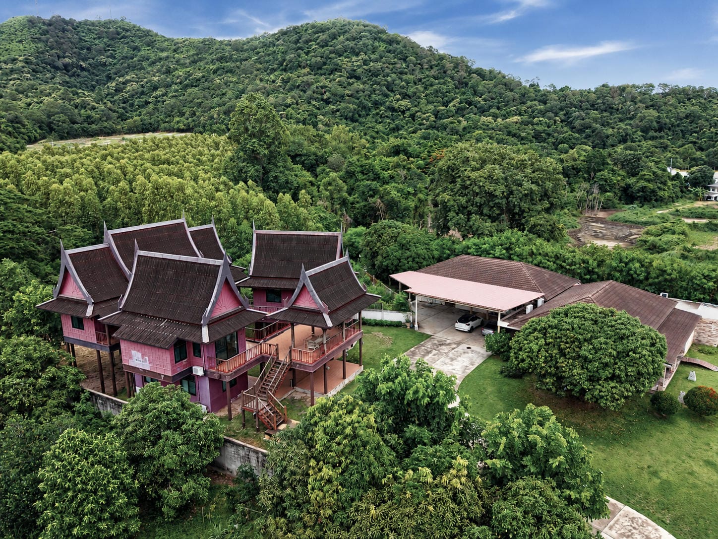 Aerial drone view of a pink two-storey Thai-style house with multiple steep gabled roofs and an external staircase, next to a single-storey building with a large carport, set in lawns surrounded by densely forested hills.