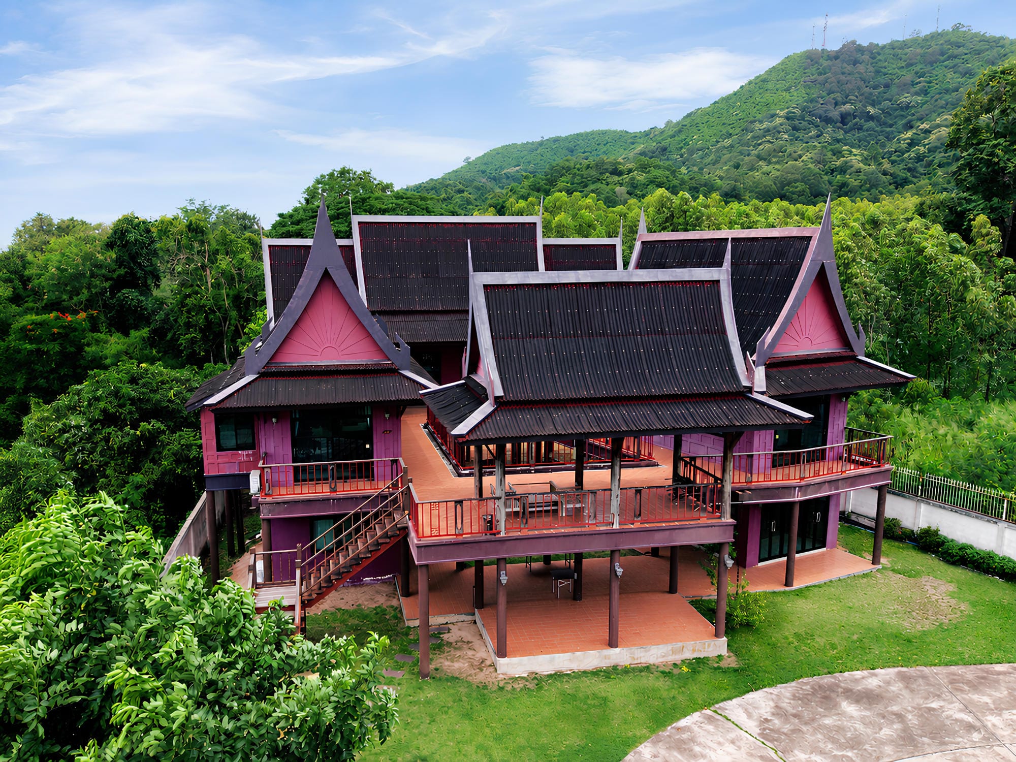 Traditional Thai-style house with steep gabled roofs and wraparound balconies on a forested hillside.