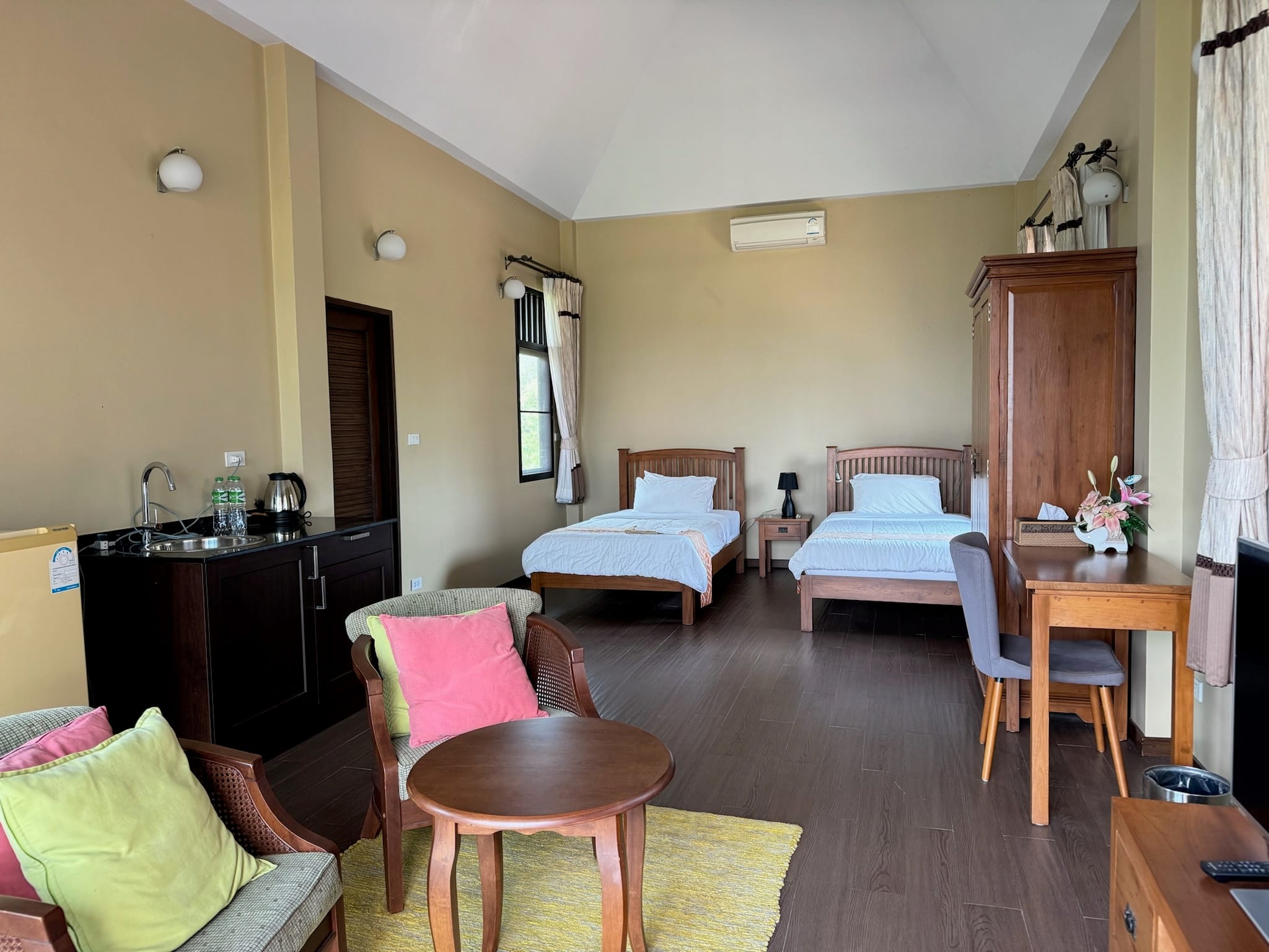 Spacious guest bedroom with a vaulted white ceiling, two wooden single beds with white linen, a dark kitchenette counter with sink and kettle, and a teak wardrobe.
