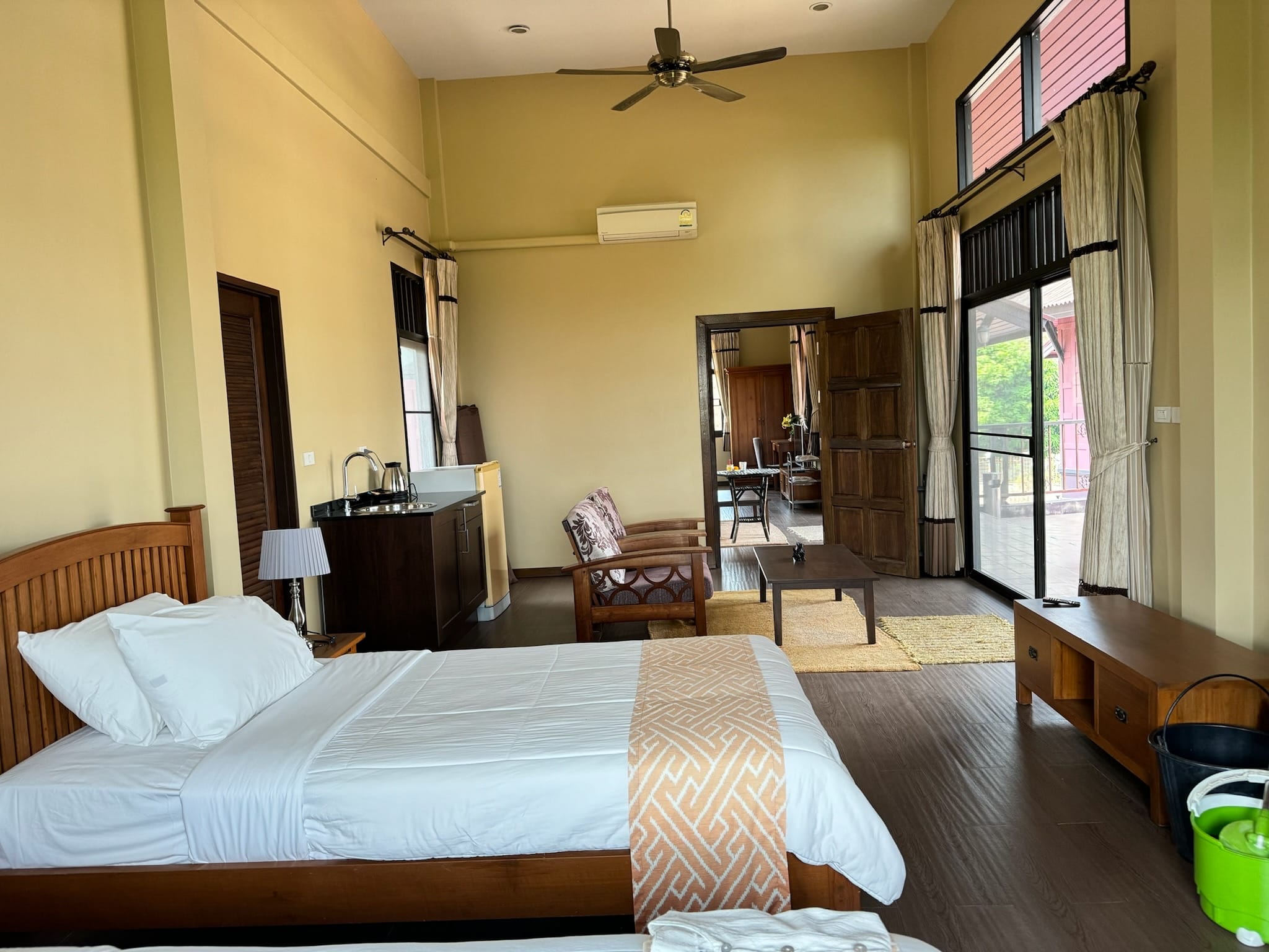 Tall-ceilinged guest bedroom with a ceiling fan, a wooden single bed with white bedding and orange patterned runner, and a dark kitchenette counter with kettle.
