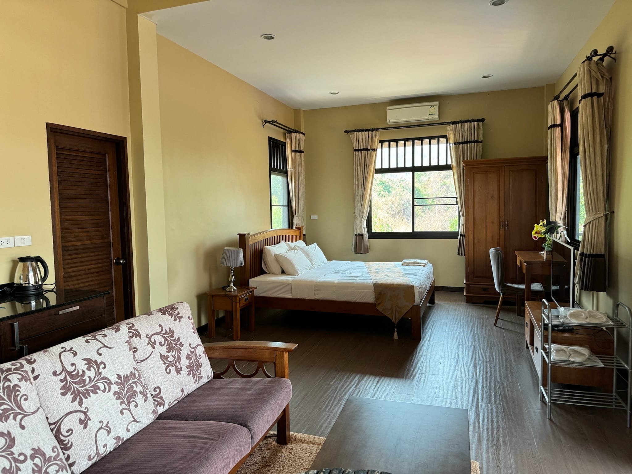 Guest bedroom with a wooden double bed dressed in white with a gold runner, a floral-upholstered wooden sofa in the foreground, and a teak wardrobe.