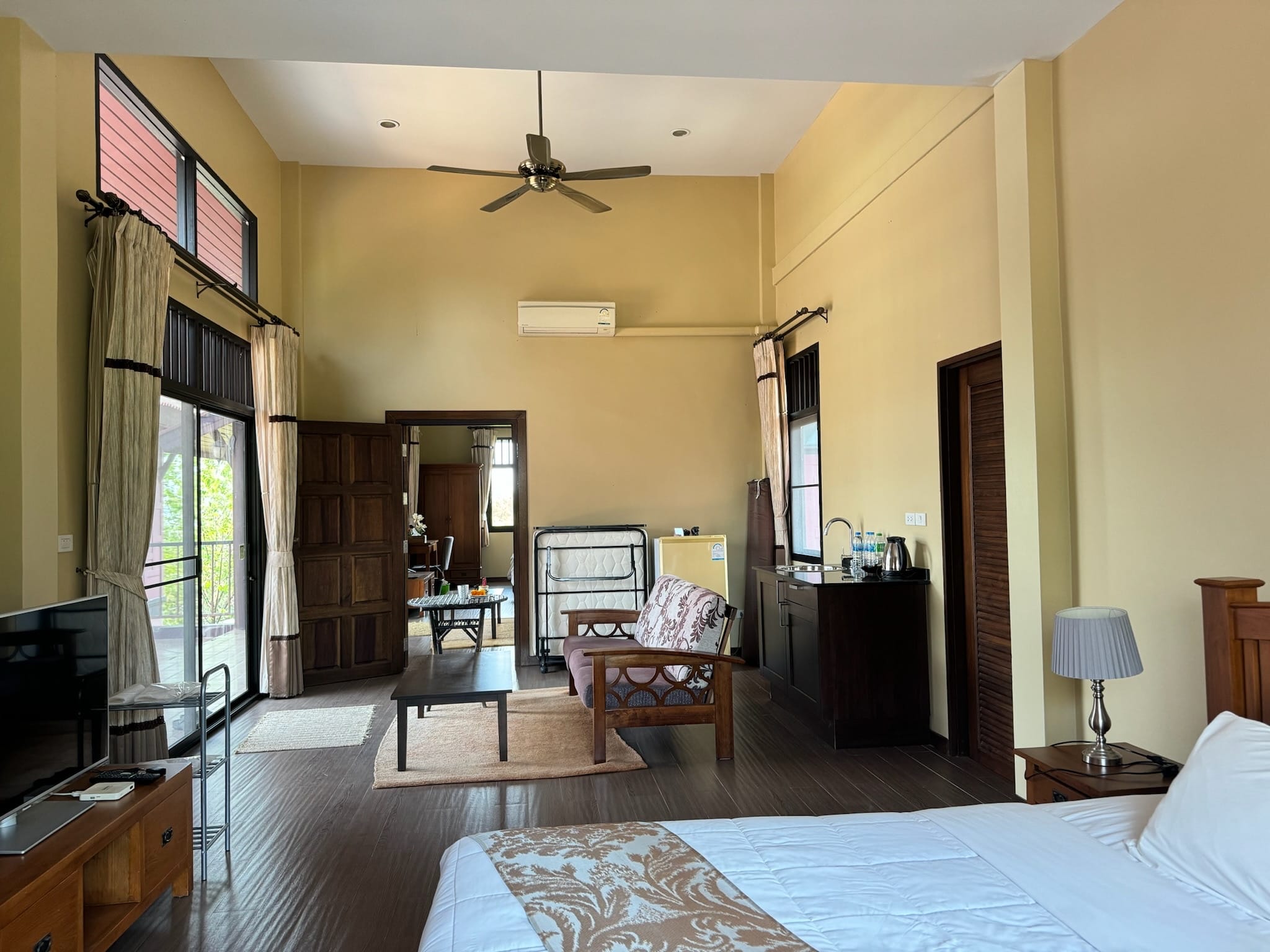 High-ceilinged suite sitting area with ceiling fan, wooden sofa with floral cushions, coffee table on a rug, and a dark kitchenette with sink and kettle.