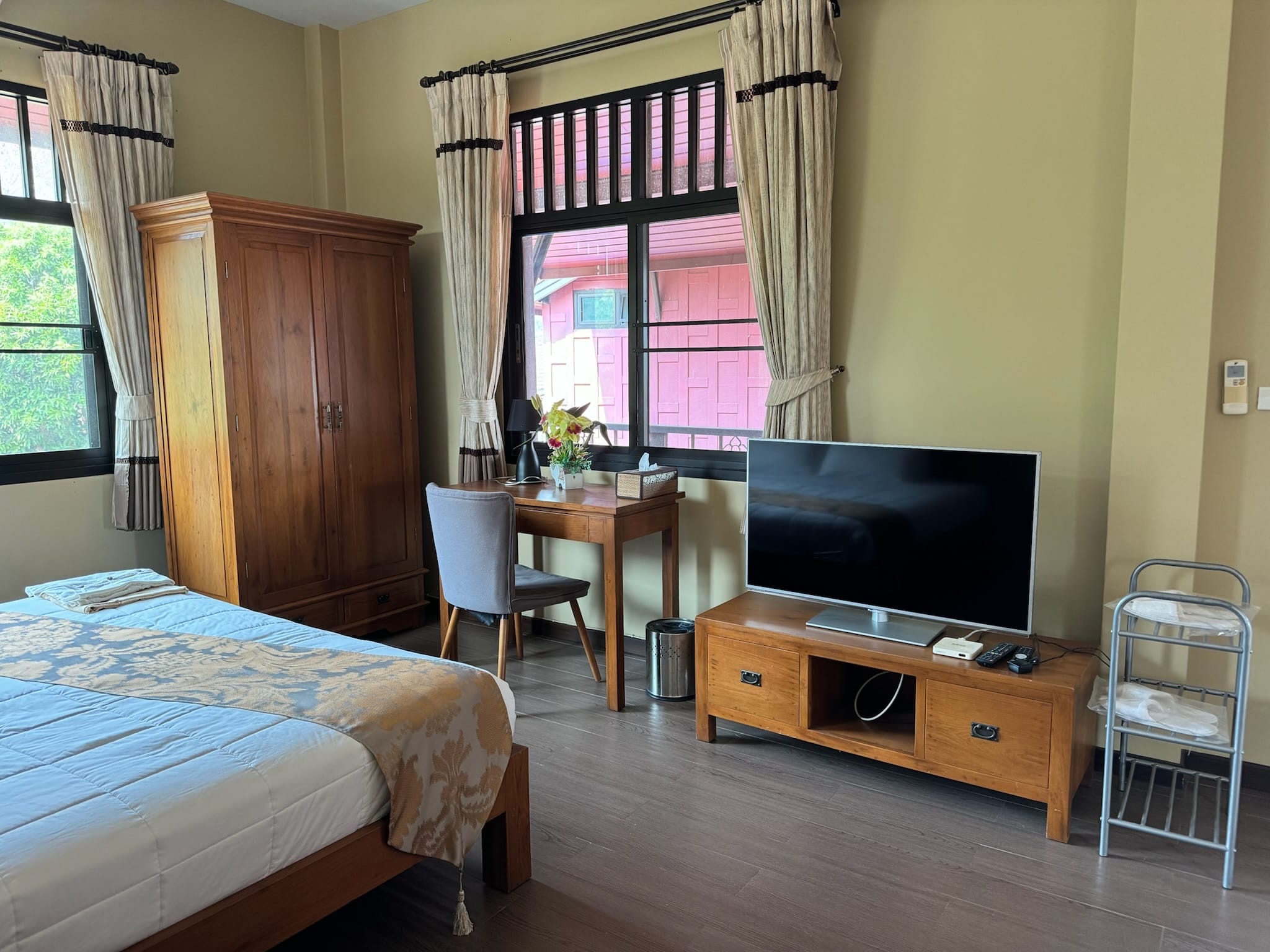 Corner of a guest bedroom showing a bed with a blue-grey damask runner, tall teak wardrobe, small desk with grey chair and flower vase, and a flat-screen TV.
