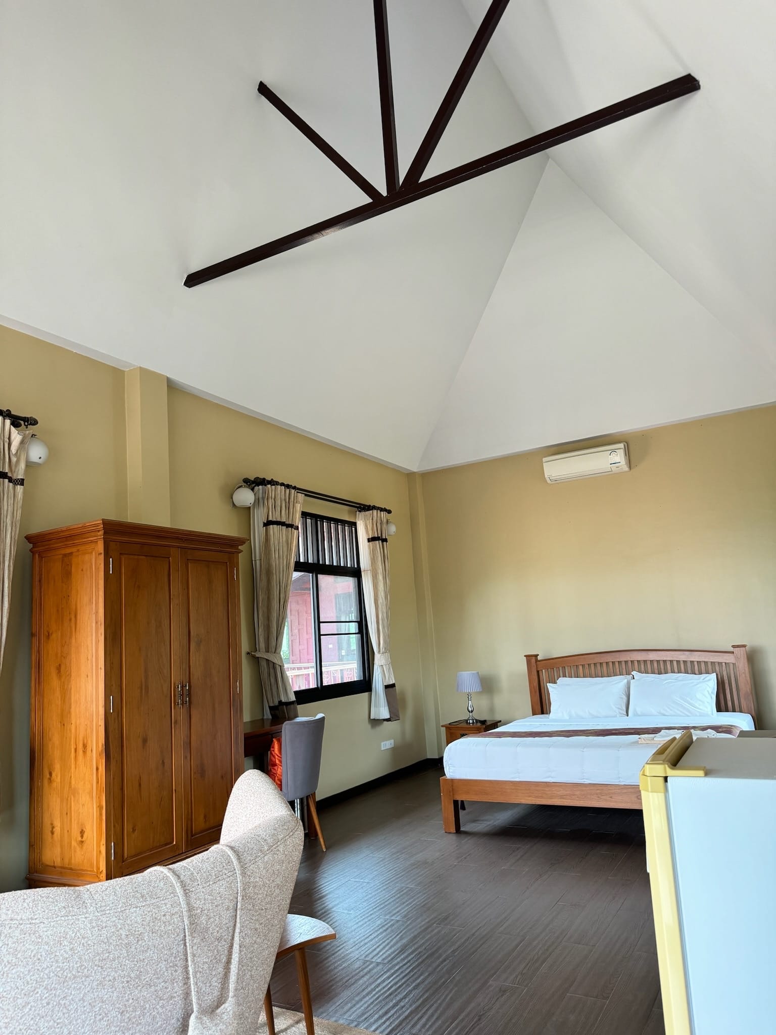 Guest bedroom interior with a high vaulted white ceiling crossed by dark timber braces, pale yellow walls, a wooden double bed with white linens, and a tall wooden wardrobe.