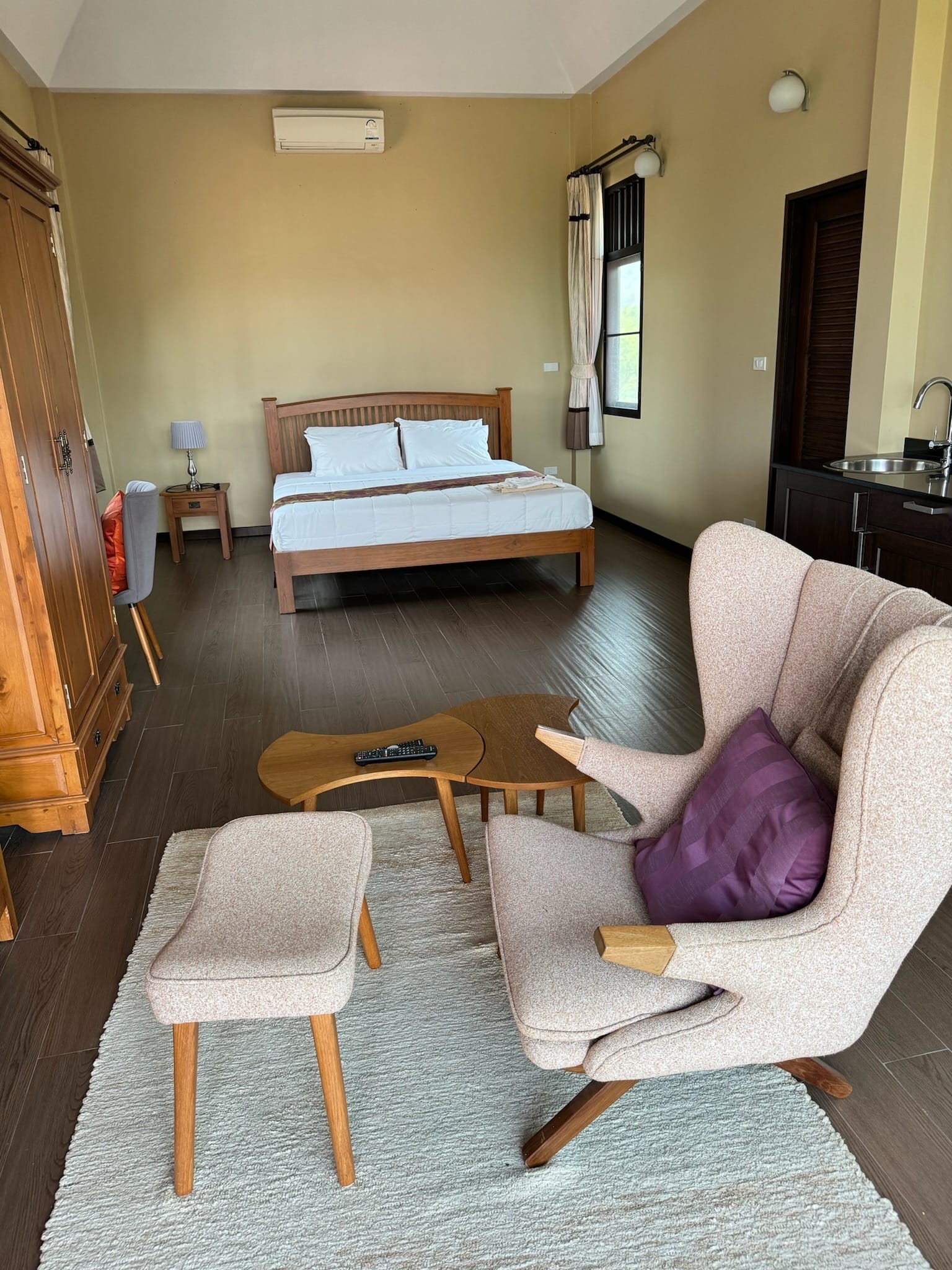 Large bedroom suite with a wooden double bed, dark wood-look floor, and a seating area in the foreground with a pale upholstered wing chair, footstool and rug.