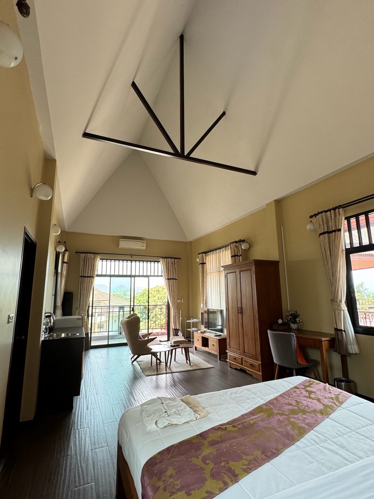 Tall vaulted bedroom suite seen from the bed, with dark timber ceiling braces, a glass balcony door opening to a terrace with hills and treetops beyond.