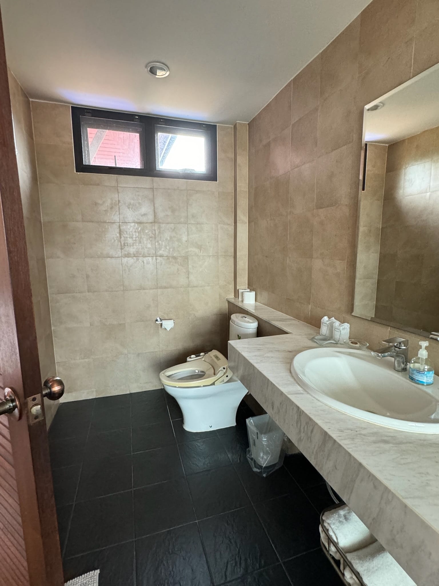 Bathroom with beige stone-look wall tiles, black tiled floor, a marble vanity counter with inset basin and large mirror, and a toilet with an electronic seat.