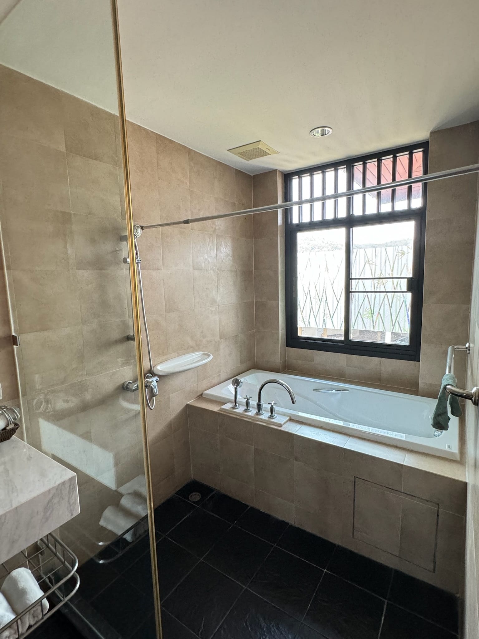 Bathroom corner with a built-in white bathtub under a barred window, chrome tub fittings, a glass shower partition with handheld shower set, and beige wall tiles.