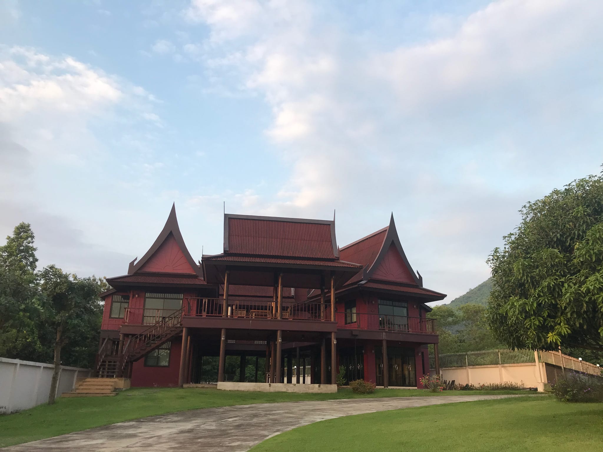 The dark red Thai house on its lawn in soft evening light, forested hill behind.